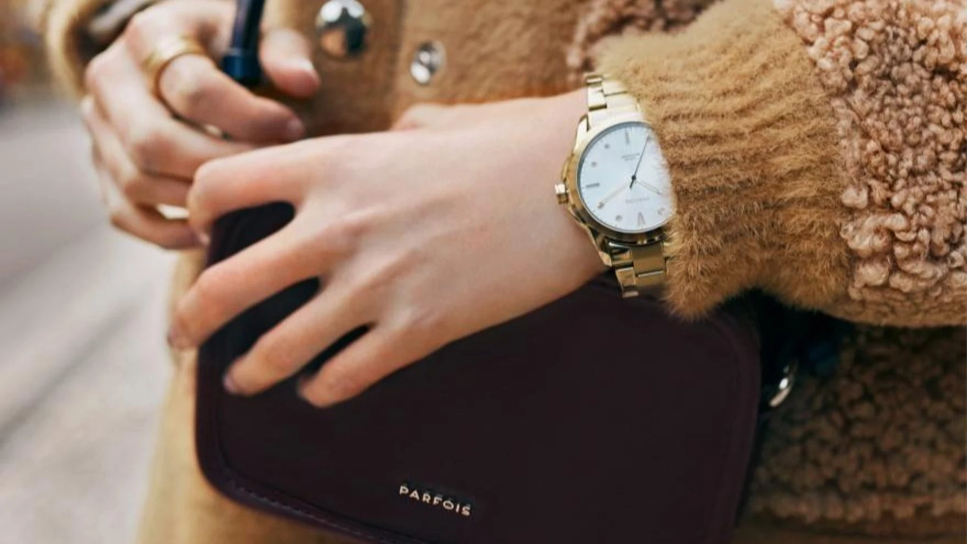 Primer plano de un reloj dorado de Parfois combinado con abrigo camel y bolso burdeos
