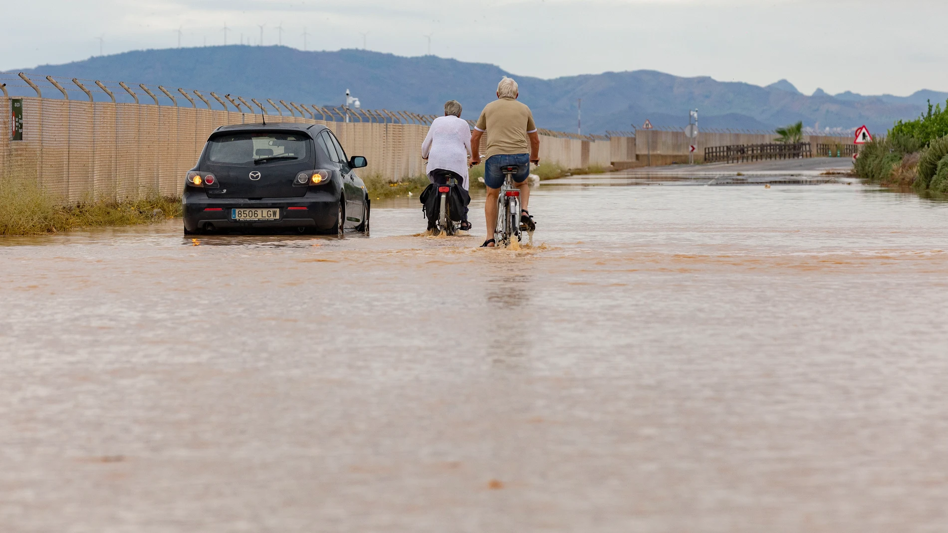 SAN JAVIER, MURCIA, 11/10/2025.- Dos ciclistas circulan junto a un coche que ha quedado atrapado por el agua en la carretera RM F-34 de San Javier (Murcia), que ha sido cortada al tráfico tras las intensas lluvias caídas la pasada noche en la comarca del mar Menor. La Comunidad Autónoma de Murcia, atendiendo a la petición del alcalde de Los Alcázares, Mario Pérez, ha comunicado que ya no es necesaria la presencia de la Unidad Militar de Emergencias en la población. El presidente de Murcia, Fe...