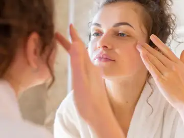 Productos para el cuidado facial de la mujer o cuidado del pelo, entre los más demandados Mujer aplicando crema facial frente al espejo, símbolo de una rutina diaria con vitaminas que fortalecen y equilibran la piel.