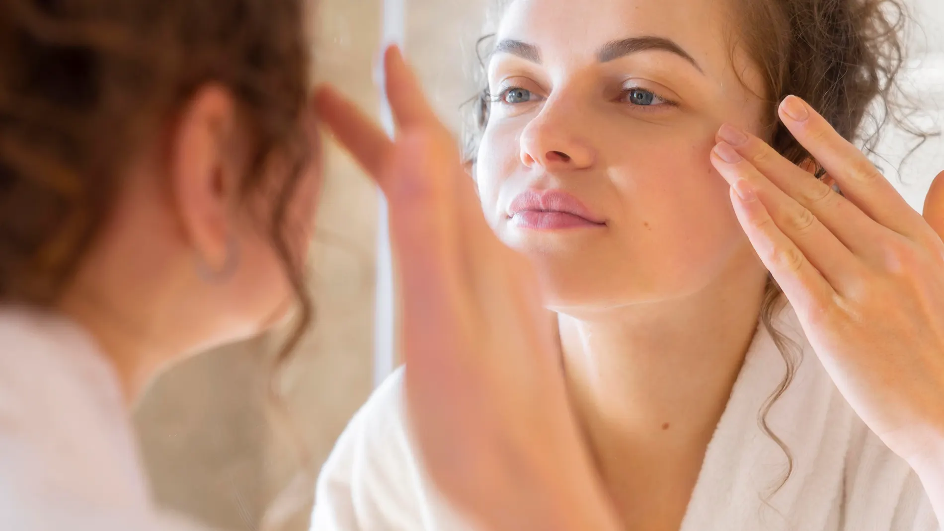 Mujer aplicando crema facial frente al espejo, símbolo de una rutina diaria con vitaminas que fortalecen y equilibran la piel.