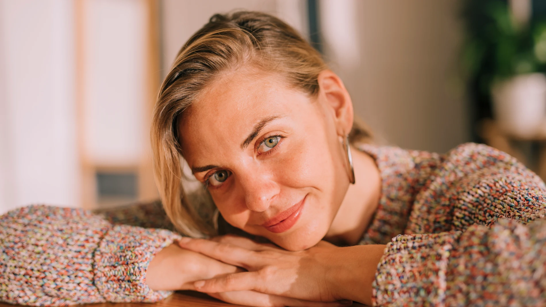 Mujer con jersey de punto apoyada sobre una mesa, sonriente y con luz cálida natural, representa una piel cuidada y saludable