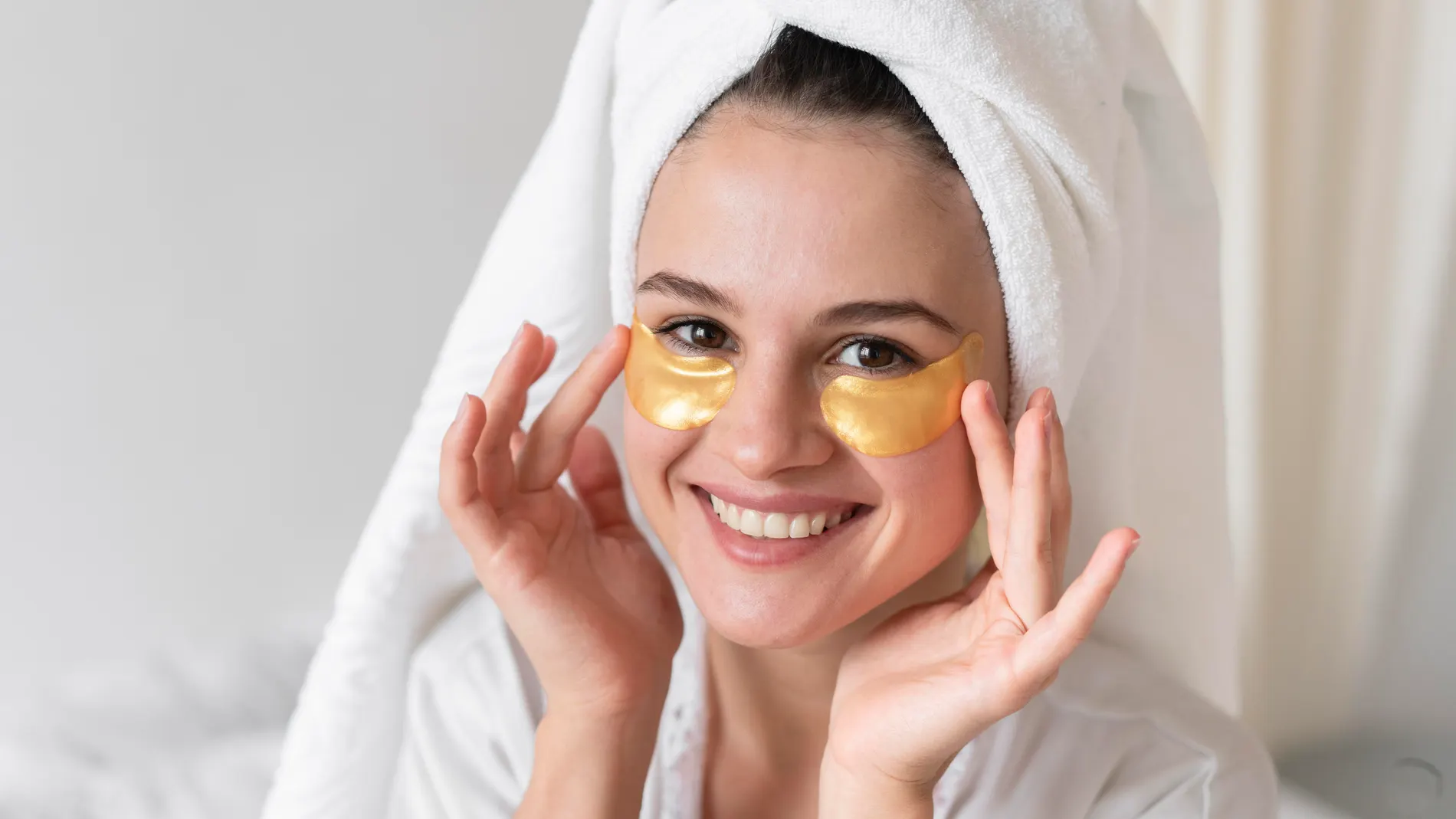 Mujer con toalla en la cabeza usando parches dorados de hidrogel en el contorno de ojos.