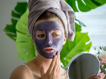 La mascarilla violeta que transforma la piel en 10 minutos: combate las rojeces y potencia la luminosidad Mujer con mascarilla facial violeta frente al espejo, durante su rutina de cuidado facial.