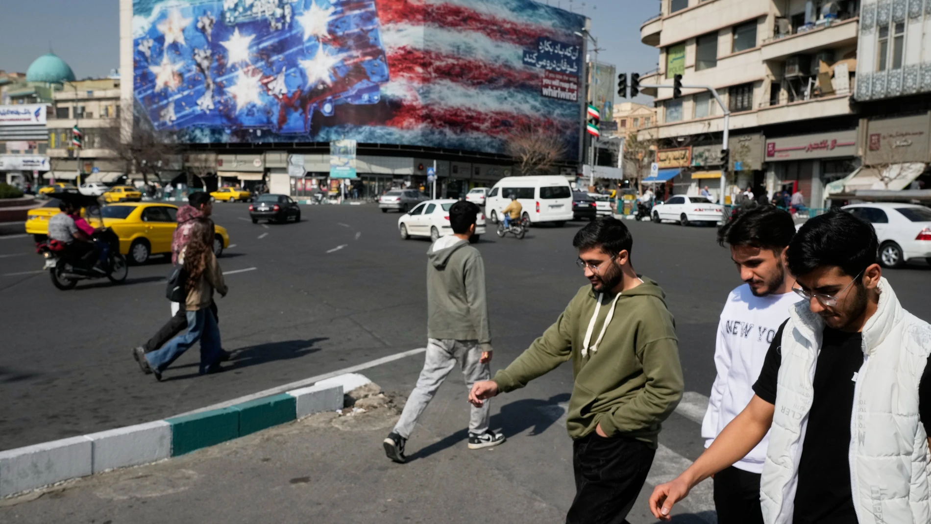 Pedestrians walk past a billboard depicting a U.S. aircraft carrier with damaged fighter jets on its deck and a sign in Farsi and English reading, "If you sow the wind, you'll reap the whirlwind," at Enqelab-e-Eslami (Islamic Revolution) Square in Tehran, Iran, Sunday, Feb. 22, 2026. (AP Photo/Vahid Salemi)